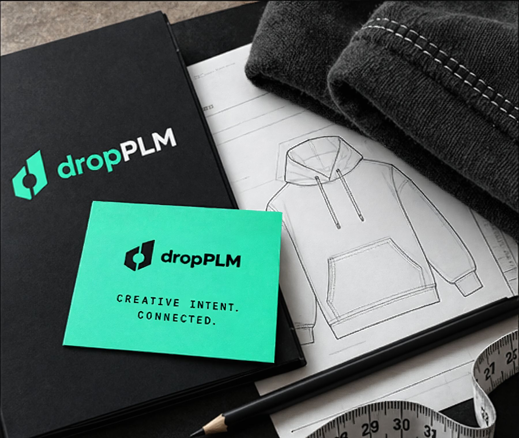 A black dropPLM notebook and a mint card reading Creative intent. Connected., on a hoodie flat sketch beside a pencil, a tape measure and a black knit